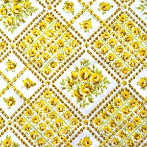 Op de afbeelding: Een witte stof met een herhalend patroon van gele rozen en groene bladeren. De rozen zijn gerangschikt in een ruitvormig patroon en worden omringd door een rand van kleine bruine stippen.