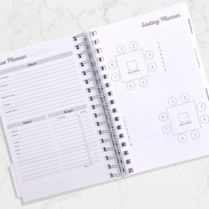 May include: Open white spiral-bound planner with sections for venue and seating arrangements. The "Venue Planner" section includes fields for details and contact information. The "Seating Planner" section features diagrams of tables with numbered seating.