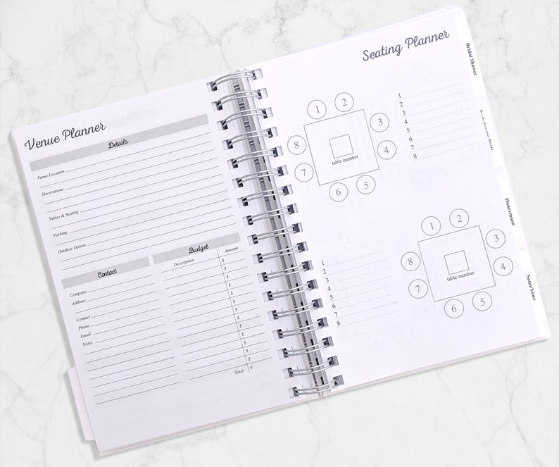 May include: Open white spiral-bound planner with sections for venue and seating arrangements. The "Venue Planner" section includes fields for details and contact information. The "Seating Planner" section features diagrams of tables with numbered seating.