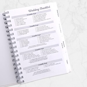 May include: A spiral-bound wedding checklist notebook with a white cover and pages. The notebook is open, displaying a detailed checklist with sections for 12, 9, 6, 5, and 4 months before the wedding. The title "Wedding Checklist" is at the top.