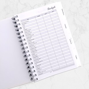 May include: Open white spiral-bound planner with a "Budget" page visible. The page is divided into columns labeled "items", "budget", "spent", and "paid". The planner is open to a checklist for a bridal shower.