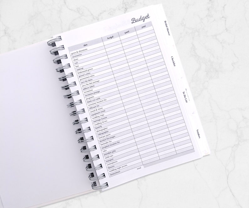 May include: Open white spiral-bound planner with a "Budget" page visible. The page is divided into columns labeled "items", "budget", "spent", and "paid". The planner is open to a checklist for a bridal shower.