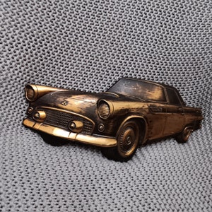 Pode incluir: Decoração de parede de carro vintage em bronze. O carro clássico apresenta um design detalhado com faróis, uma grelha e uma carroçaria elegante. O carro está sobre um fundo cinzento texturizado, criando uma estética vintage.