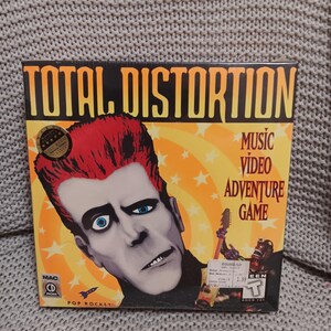 May include: A sealed Total Distortion music video adventure game box for Mac CD-ROM. The cover features a stylized illustration of a person with red hair and blue eyes against a yellow starburst background, with the title text at the top.