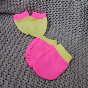 May include: Two pairs of baby mittens. The mittens are a bright neon pink with a yellow cuff. The mittens are laying on a gray knitted blanket. The mittens are made of a soft material.