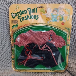 May include: A doll outfit in original packaging labelled Garden Doll Fashions, including a red and white gingham puff-sleeve top with a small sailboat graphic and dark blue shorts with red trim, accompanied by a red plastic heart-shaped hanger.