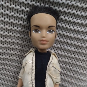 Könnte beinhalten: Eine Puppe mit schwarzen Haaren, brauner Haut und blauen Augen. Die Puppe trägt ein weißes Hemd mit Knöpfen und schwarzem Besatz und darunter ein schwarzes T-Shirt.