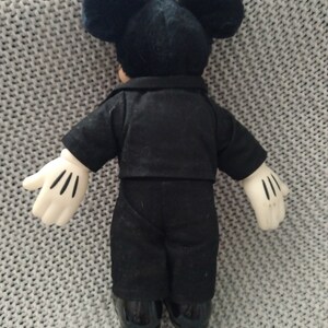 May include: A black plush Mickey Mouse doll wearing a black suit and white gloves. The doll is standing on a gray knitted surface.