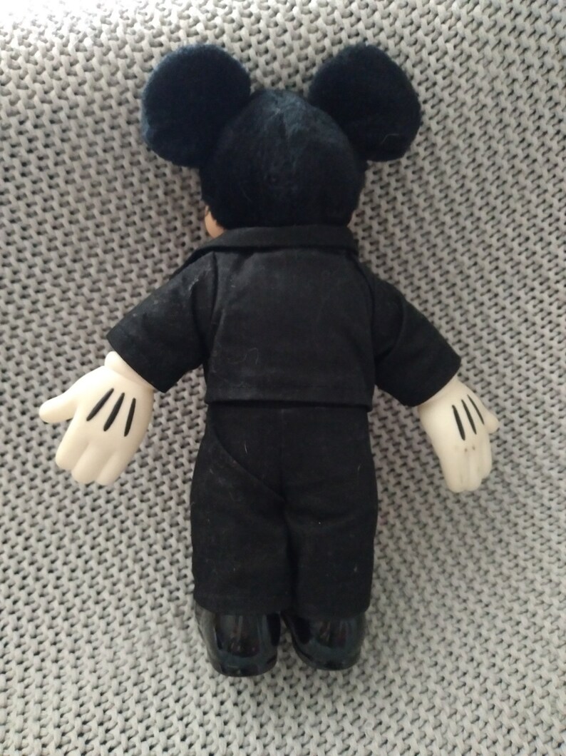 May include: A black plush Mickey Mouse doll wearing a black suit and white gloves. The doll is standing on a gray knitted surface.