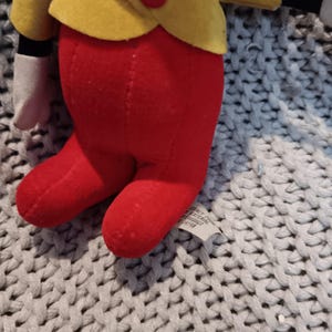 May include: A plush doll featuring a yellow jacket with a red button, red pants, and white gloves. The doll has a blue bow tie and a red nose. The doll is made of soft fabric and is likely a children's toy.