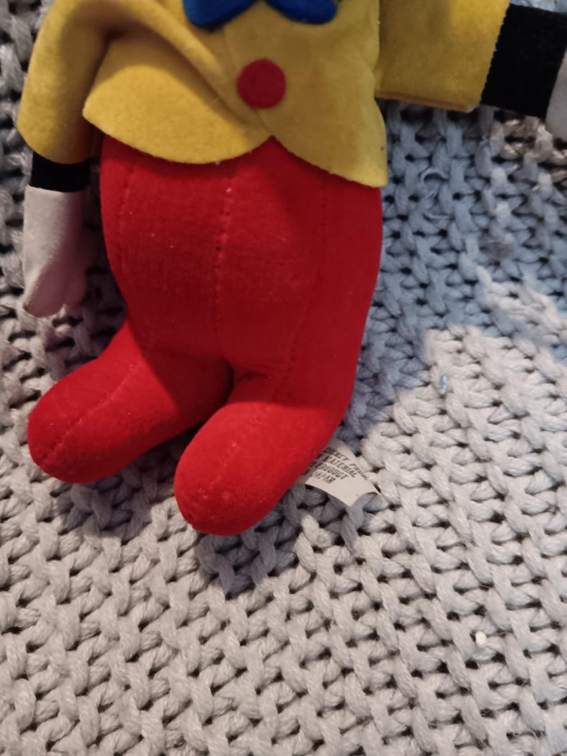 May include: A plush doll featuring a yellow jacket with a red button, red pants, and white gloves. The doll has a blue bow tie and a red nose. The doll is made of soft fabric and is likely a children's toy.