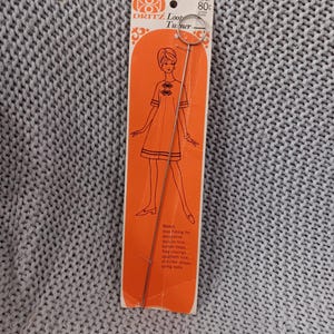 May include: An orange and white Dritz Loop Turner tool on a white and orange card. The card features a drawing of a woman in a dress. The tool is made of metal and has a loop on one end. The card has text that says "Loop Turner".