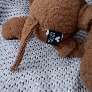 K&ouml;nnte beinhalten: Ein braunes Pl&uuml;schtier mit Schwanz und Ohren. Das Spielzeug hat ein Etikett mit der Aufschrift "SPECIAL EDITION" mit einer Mickey Mouse-Silhouette. Das Spielzeug liegt auf einer grauen Strickoberfl&auml;che.