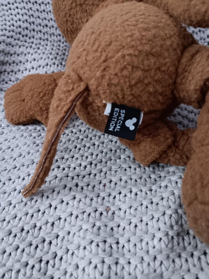 K&ouml;nnte beinhalten: Ein braunes Pl&uuml;schtier mit Schwanz und Ohren. Das Spielzeug hat ein Etikett mit der Aufschrift "SPECIAL EDITION" mit einer Mickey Mouse-Silhouette. Das Spielzeug liegt auf einer grauen Strickoberfl&auml;che.