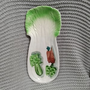 Puede incluir: Una fuente de cerámica blanca con forma de hoja de lechuga, con colores verdes y blancos. La fuente está decorada con imágenes pintadas de brócoli, zanahorias y otras verduras.