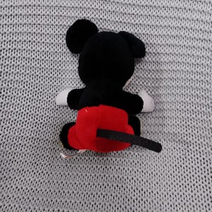 K&ouml;nnte beinhalten: Ein schwarzes und rotes Pl&uuml;sch-Mickey-Mouse-Spielzeug mit einem schwarzen Schwanz. Das Spielzeug sitzt auf einer grauen gestrickten Oberfl&auml;che.