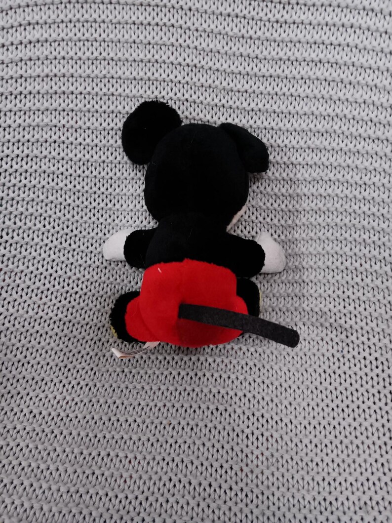 K&ouml;nnte beinhalten: Ein schwarzes und rotes Pl&uuml;sch-Mickey-Mouse-Spielzeug mit einem schwarzen Schwanz. Das Spielzeug sitzt auf einer grauen gestrickten Oberfl&auml;che.