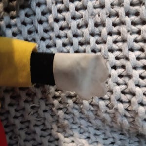 May include: A close-up of a plush toy's hand. The hand is white with a rounded shape. The sleeve is yellow with a black cuff. The background is a gray, textured fabric.