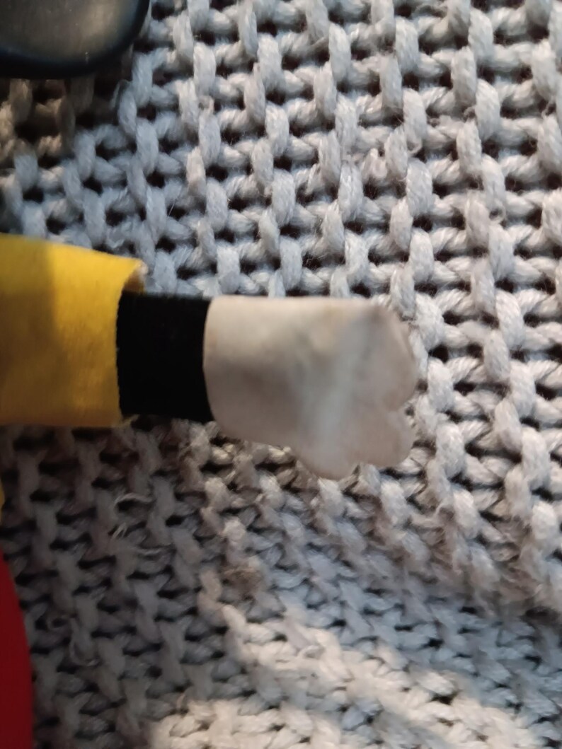 May include: A close-up of a plush toy's hand. The hand is white with a rounded shape. The sleeve is yellow with a black cuff. The background is a gray, textured fabric.