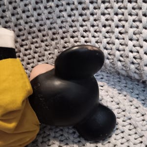 May include: A black Mickey Mouse ear with a pink inner ear and a yellow garment. The ear is made of a smooth, shiny material. The background is a gray knitted fabric.
