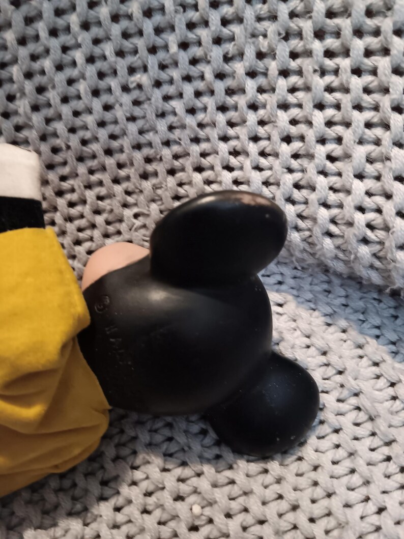 May include: A black Mickey Mouse ear with a pink inner ear and a yellow garment. The ear is made of a smooth, shiny material. The background is a gray knitted fabric.