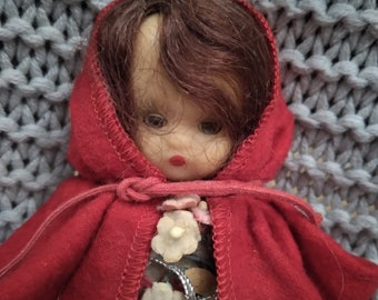 Puede incluir: Una muñeca pequeña con una capa roja con capucha y flores blancas en la parte delantera. La muñeca tiene el pelo castaño y la cara pintada.