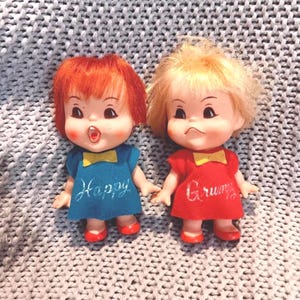 May include: Two vintage dolls with expressive faces. One doll wears a blue dress with the word "Happy," and the other wears a red dress with the word "Grumpy." Both dolls have red shoes and yellow bow ties.