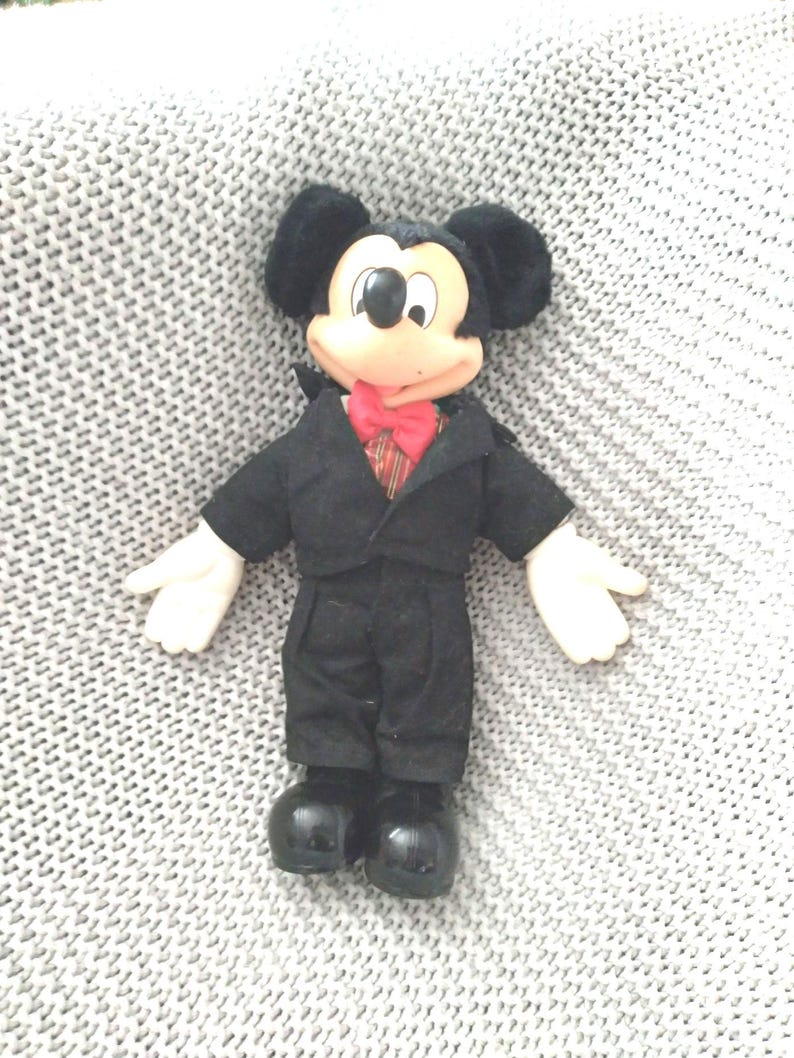 May include: A plush Mickey Mouse doll wearing a black tuxedo with a red bow tie. The doll has a black nose and large black ears.