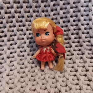 May include: A small plastic doll with blonde hair and blue eyes. The doll is wearing a red dress and has a red bow in her hair. The doll is resting on a gray knitted surface.