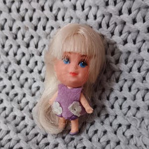 May include: A small doll with blonde hair and blue eyes. The doll wears a purple dress with white flower accents. The doll is approximately 2 inches tall. The background is a gray knitted fabric.