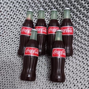 May include: Six miniature Coca-Cola bottles with silver caps. The bottles are brown and have the Coca-Cola logo in red and white.