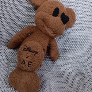 K&ouml;nnte beinhalten: Ein braunes Mickey Mouse Pl&uuml;schtier mit schwarzen Augen und Nase. Das Spielzeug hat das Disney x AE Logo auf der R&uuml;ckseite. Das Pl&uuml;schtier liegt auf einer grauen Strickoberfl&auml;che.