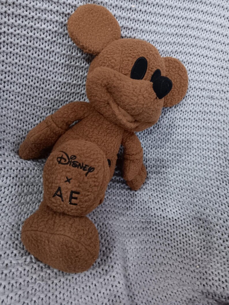 K&ouml;nnte beinhalten: Ein braunes Mickey Mouse Pl&uuml;schtier mit schwarzen Augen und Nase. Das Spielzeug hat das Disney x AE Logo auf der R&uuml;ckseite. Das Pl&uuml;schtier liegt auf einer grauen Strickoberfl&auml;che.