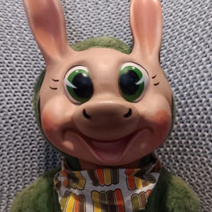Puede incluir: Un juguete de peluche vintage con un diseño caprichoso. El juguete presenta una cara sonriente con grandes ojos verdes, mejillas rosadas y largas orejas rosas. Viste un atuendo verde y un pañuelo estampado. El juguete tiene una apariencia alegre y caricaturesca.