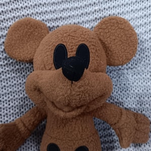 K&ouml;nnte beinhalten: Ein braunes Pl&uuml;schtier in Form einer Zeichentrickmaus mit gro&szlig;en, runden Ohren. Die Maus hat schwarze Augen, eine schwarze Nase und zwei schwarze Kreise auf ihrem K&ouml;rper. Das Spielzeug besteht aus einem weichen, flauschigen Material.