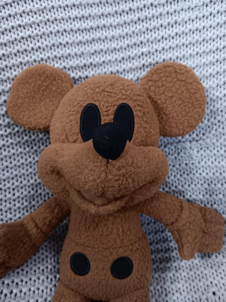 K&ouml;nnte beinhalten: Ein braunes Pl&uuml;schtier in Form einer Zeichentrickmaus mit gro&szlig;en, runden Ohren. Die Maus hat schwarze Augen, eine schwarze Nase und zwei schwarze Kreise auf ihrem K&ouml;rper. Das Spielzeug besteht aus einem weichen, flauschigen Material.