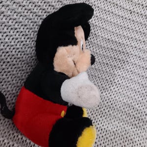 K&ouml;nnte beinhalten: Ein Pl&uuml;sch-Mickey-Mouse-Spielzeug, das auf einer grauen Strickoberfl&auml;che sitzt. Das Spielzeug ist schwarz, rot und gelb. Mickey Mouse tr&auml;gt seine charakteristischen roten Shorts und gelben Schuhe.