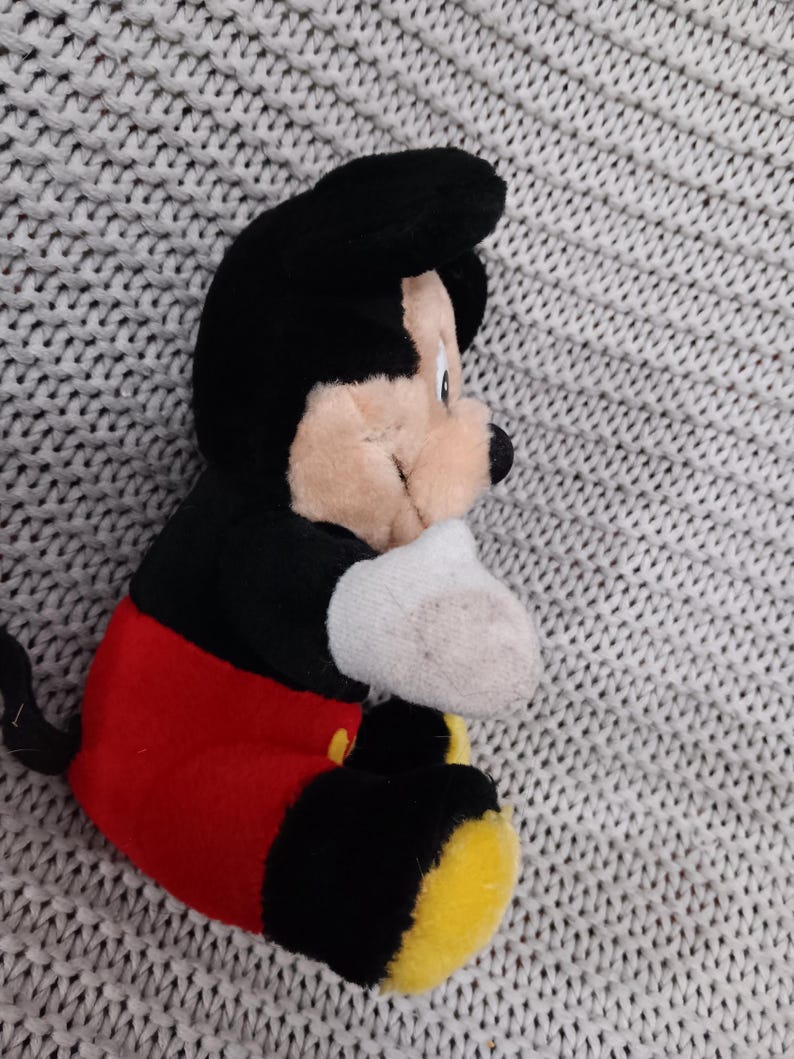 K&ouml;nnte beinhalten: Ein Pl&uuml;sch-Mickey-Mouse-Spielzeug, das auf einer grauen Strickoberfl&auml;che sitzt. Das Spielzeug ist schwarz, rot und gelb. Mickey Mouse tr&auml;gt seine charakteristischen roten Shorts und gelben Schuhe.