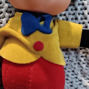 May include: A vintage plush toy featuring a smiling face with a black nose and ears. The toy is dressed in a yellow jacket with a blue bow tie and red pants. A red button accents the jacket. The toy is a classic character.