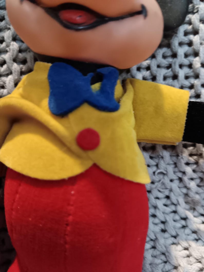 May include: A vintage plush toy featuring a smiling face with a black nose and ears. The toy is dressed in a yellow jacket with a blue bow tie and red pants. A red button accents the jacket. The toy is a classic character.