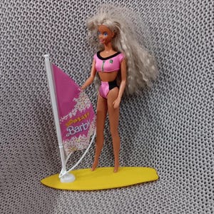 May include: A vintage Barbie doll in a pink and black swimsuit, holding a pink and white windsurfing sail. The doll stands on a yellow surfboard. The sail reads "Surf Blast Barbie".