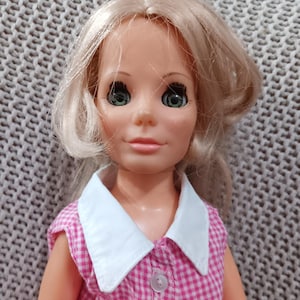 May include: A vintage doll with blonde hair, green eyes, and a pink and white gingham dress. The doll has a white collar and a button on the front of the dress.