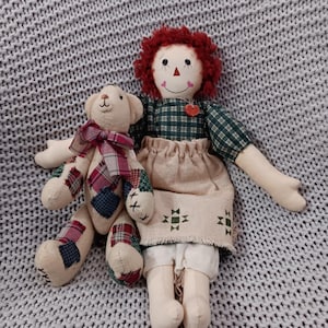 Könnte beinhalten: Eine handgefertigte Stoffpuppe mit roten Garnhaaren, einem grün karierten Oberteil und einem beigen Rock, die einen Patchwork-Teddybären mit einer karierten Schleife hält. Die Puppe hat ein gemaltes Gesicht und ein herzförmiges Detail.