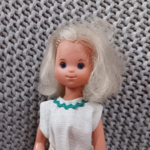 May include: A vintage doll with blonde hair and blue eyes. The doll wears a white dress with a green scalloped neckline. The doll is made of plastic and has a neutral expression.