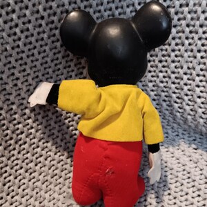 May include: A vintage Mickey Mouse doll, viewed from the back, with a black head and ears, a yellow jacket, red pants, and white gloves. The doll is posed with one arm extended. The background is a gray knitted fabric.