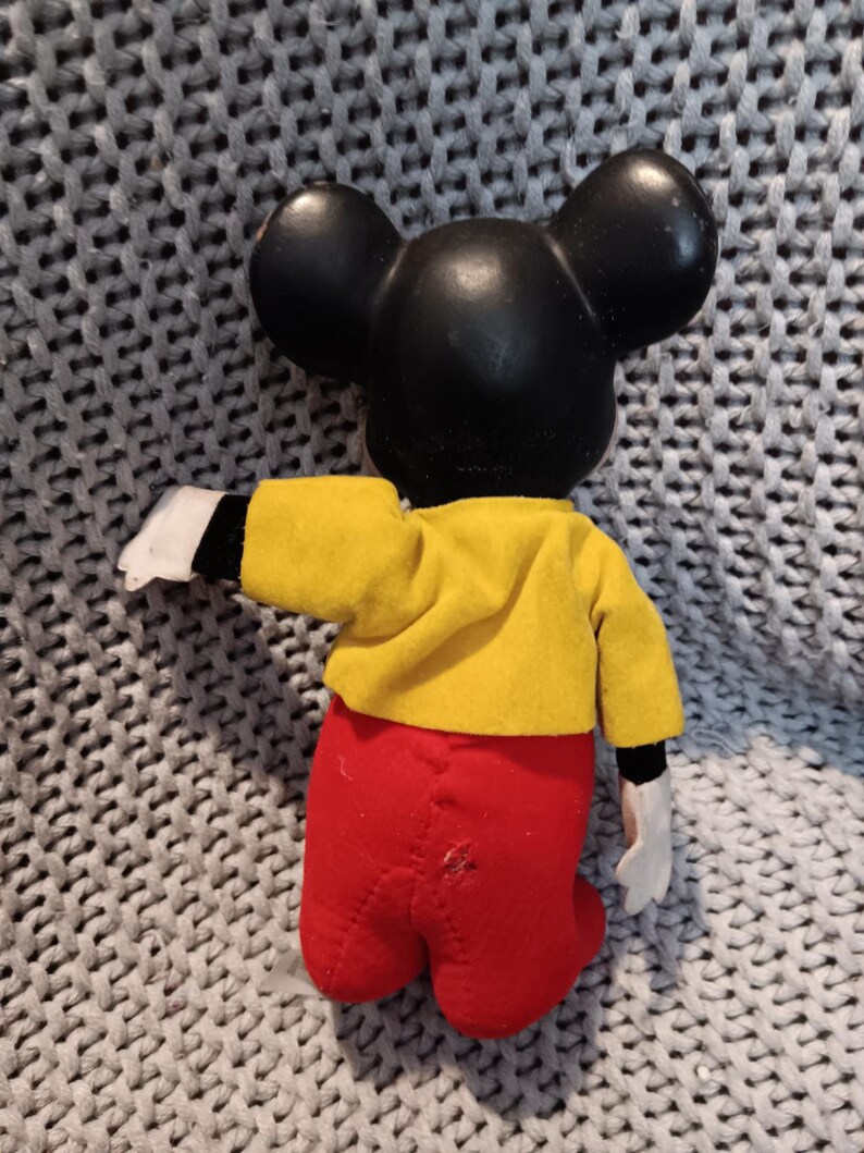 May include: A vintage Mickey Mouse doll, viewed from the back, with a black head and ears, a yellow jacket, red pants, and white gloves. The doll is posed with one arm extended. The background is a gray knitted fabric.