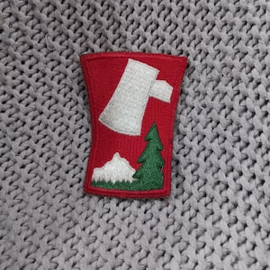 Può includere: Una toppa rossa ricamata con un'ascia bianca, un pino verde e una catena montuosa bianca. La toppa è rettangolare con angoli arrotondati, su uno sfondo a maglia grigio.
