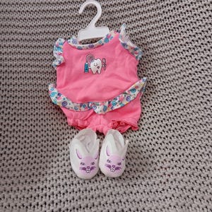 May include: A pink doll outfit with a tooth and toothbrush design. The outfit has a ruffled skirt and comes with a pair of white bunny slippers.