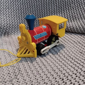 May include: A vintage toy train in yellow, red, and blue. The train has the words "TOOT-TOOT" printed on the side. The train has black wheels and a yellow pull string.
