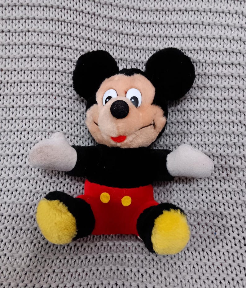 K&ouml;nnte beinhalten: Ein schwarzes und rotes Pl&uuml;sch-Mickey-Mouse-Spielzeug mit gelben Schuhen und einem breiten L&auml;cheln. Das Spielzeug sitzt auf einer grauen gestrickten Oberfl&auml;che.